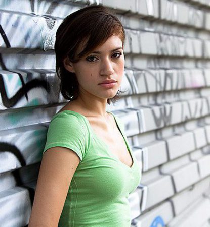 A teenage girl stands up against a metal barrier covered in graffiti, that extends all the way across the picture, fading out to a blur as it gets farther away.