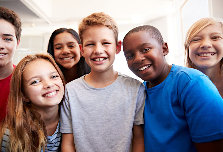 A close-up group portrait of six smiling children standing in a bright indoor setting. They appear to be about 10 to 12 years old, and vary in race, with different skin tones and hair colors. They are dressed casually in t-shirts and are positioned close together, filling the frame. The focus is on their cheerful faces as they look toward the camera.