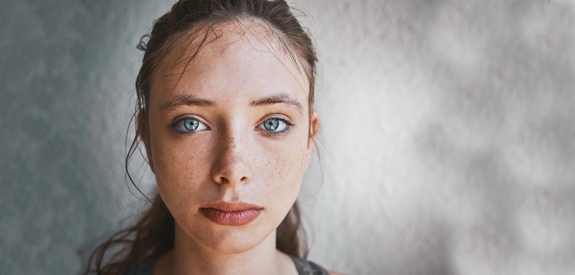 Close-up portrait of a young female facing the camera against a neutral light gray background. She has light skin with freckles, blue-gray eyes looking directly at the viewer, and straight brown hair pulled back behind her ears with a few loose strands on her forehead. Her expression is neutral and calm. The framing shows her head and upper shoulders centered in the image.