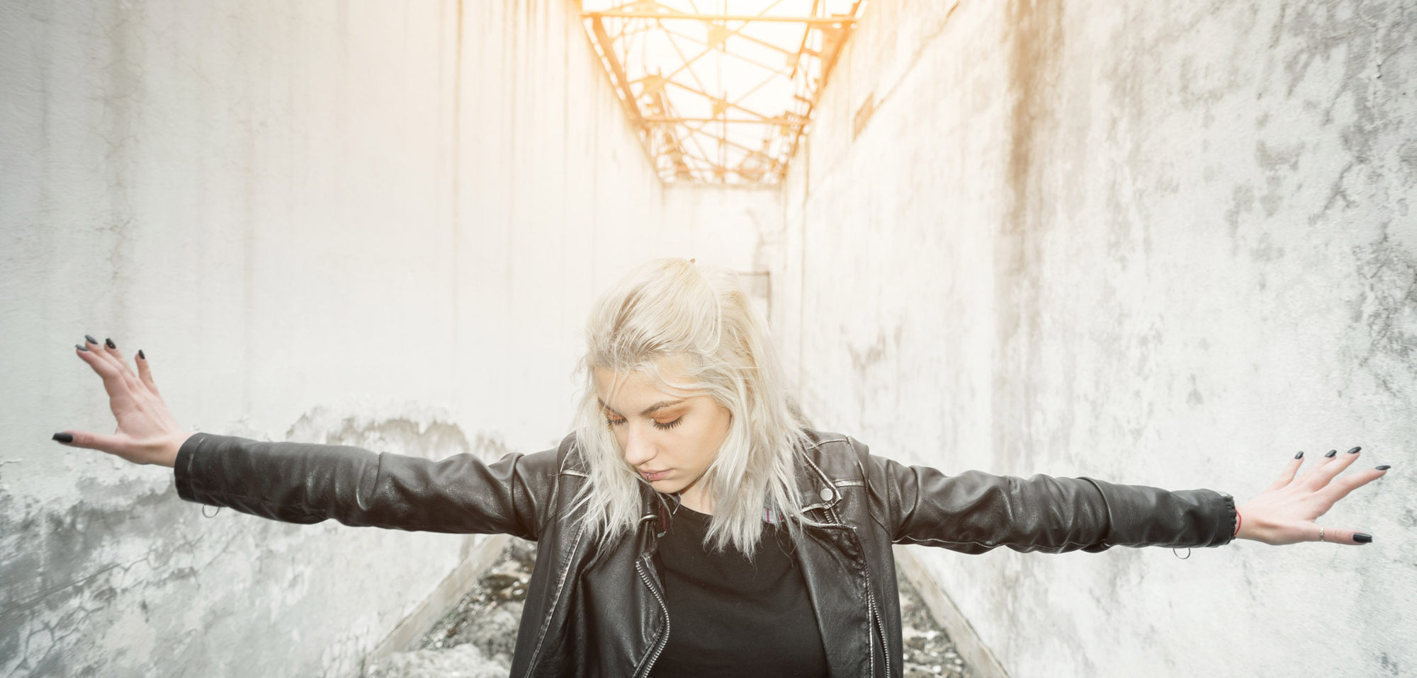 A young, white-haired girl in a black leather jacket with black fingernails has her hands out, fingers against white walls on both sides, inside a run-down industrial building.