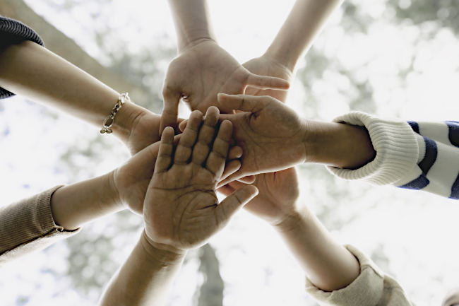 A picture of 7 hands, each from a different person, forming what resembles the spokes on a wagon wheel. Light is coming from above the hands and through blurry tree branches.