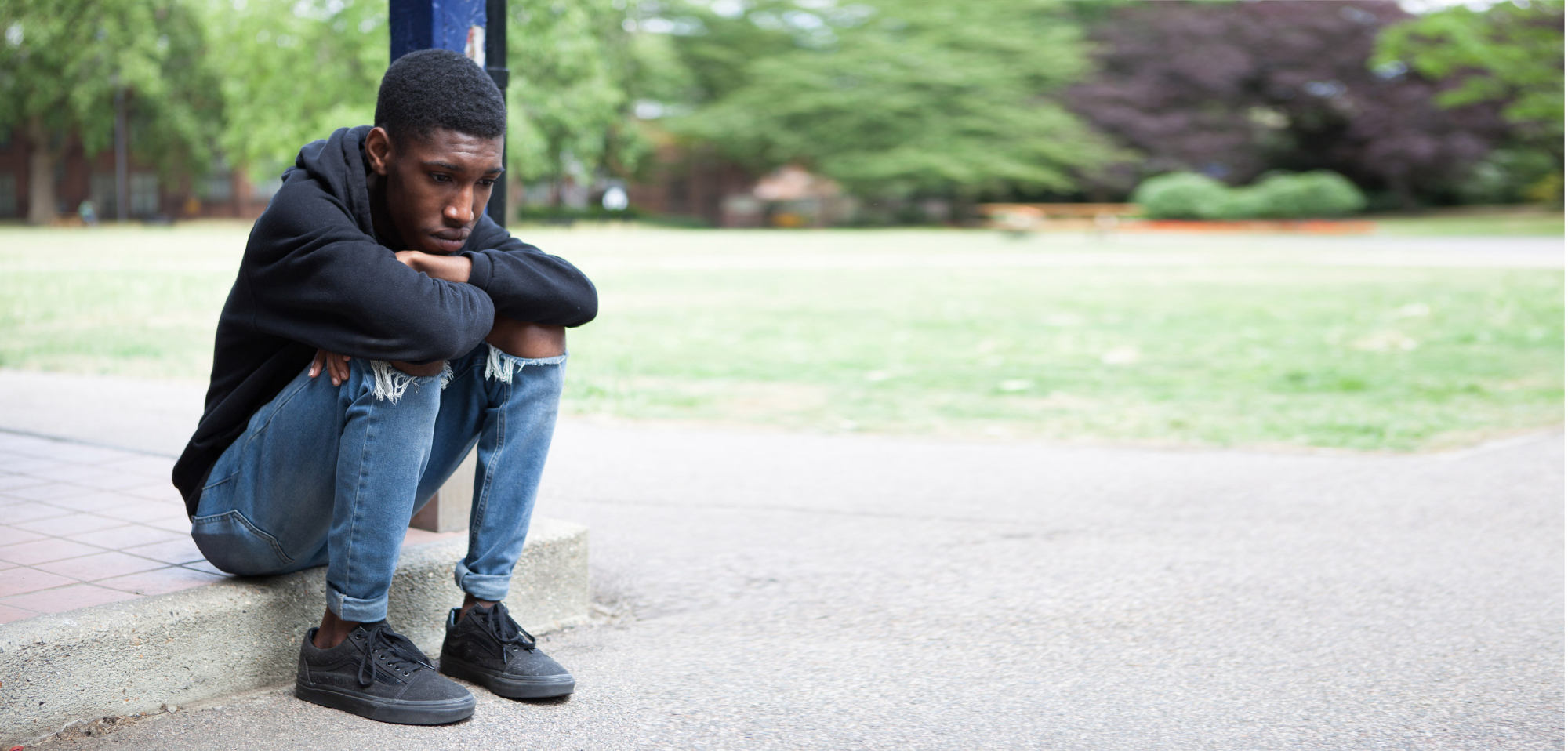 Teenage boy sitting on a low concrete step outdoors with his arms resting on his knees, wearing a black hoodie and jeans, looking downward in a thoughtful pose.