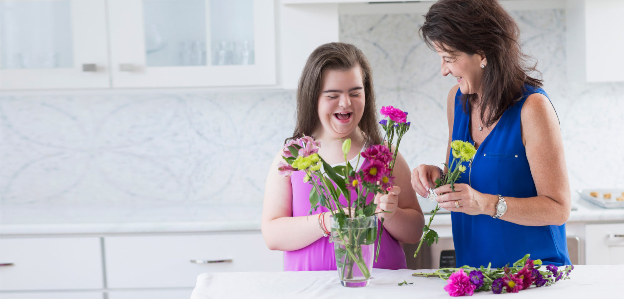 A mother is laughing with her daughter, and looking at her as her daughter looks down. They are both placing flowers in a clear glass vase on a white table in the kitchen. All the cabinets and drawers are white.