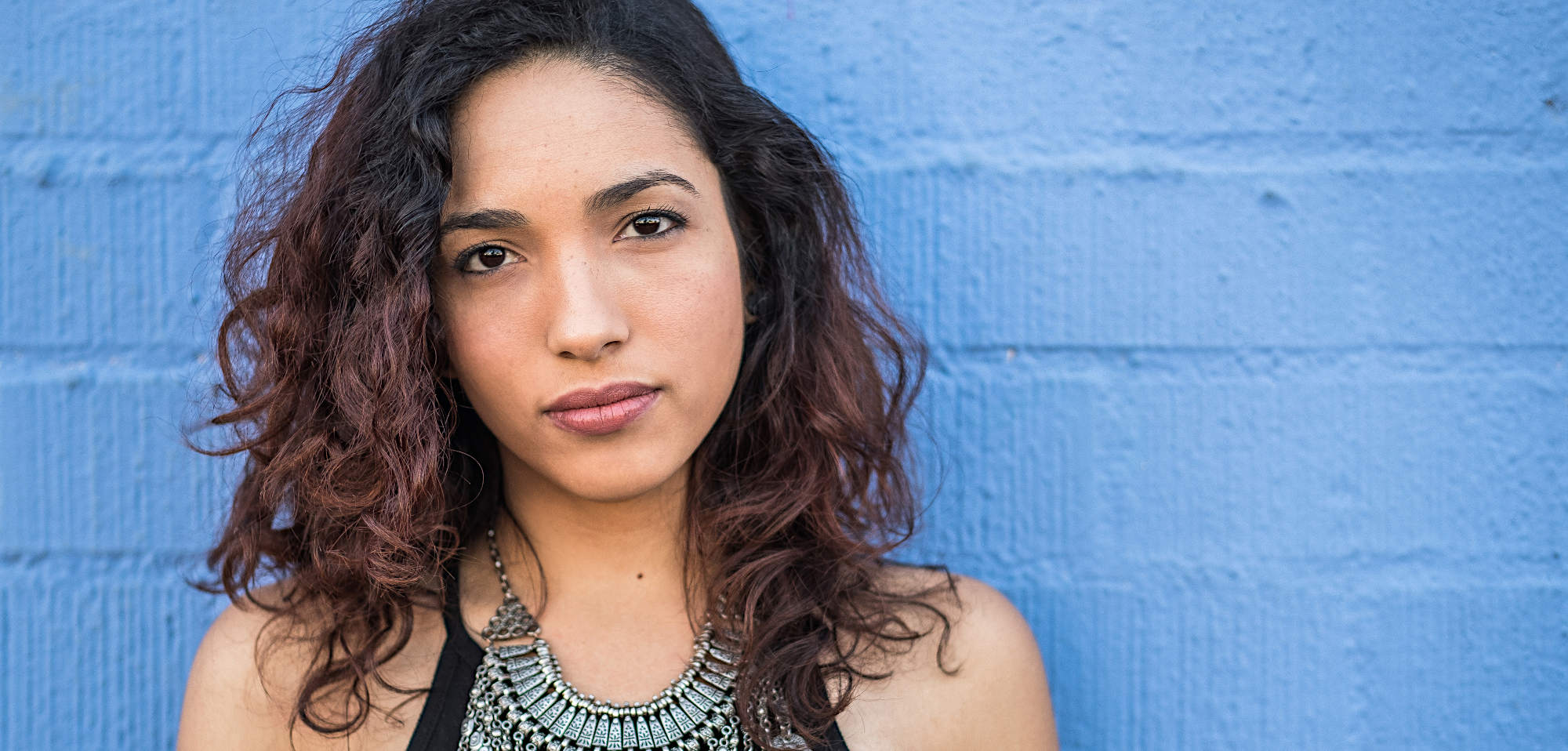 A close-up portrait of a young woman standing in front of a bright blue painted brick wall. She has medium-length, wavy dark hair with reddish highlights, warm brown skin, and a neutral, confident expression. She’s wearing a sleeveless black top and a large, ornate silver statement necklace centered on her chest. The background is blurred slightly, keeping focus on her face and upper body.